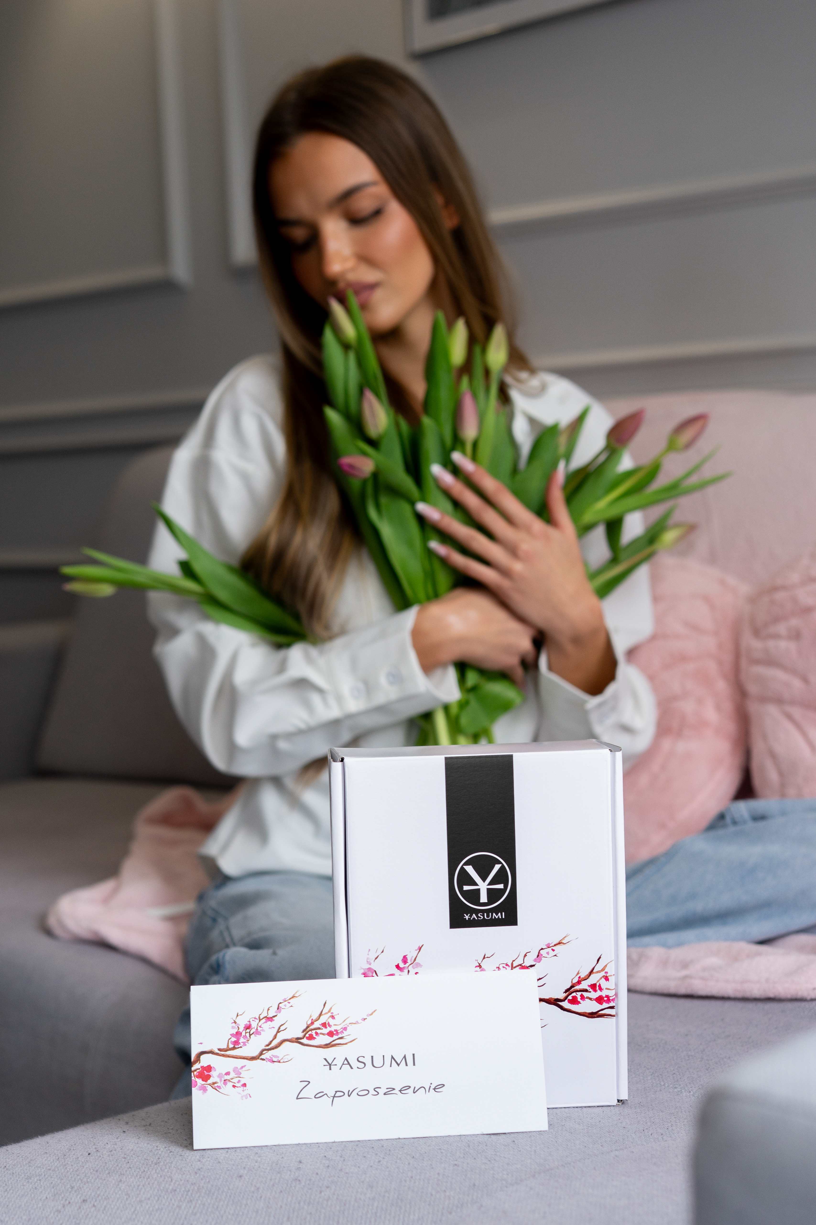 Kobieta z bukietem tulipanów, a przed nią pudełko i voucher prezentowy YASUMI